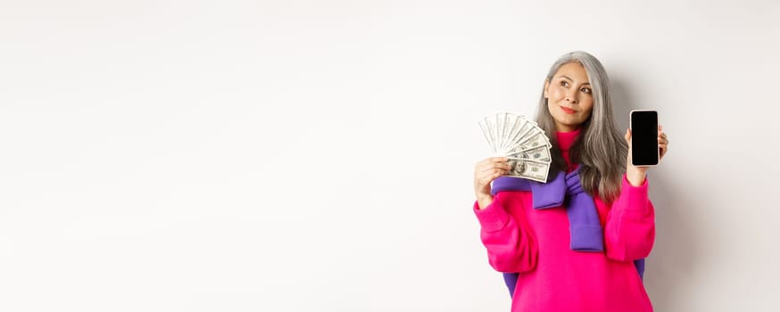 Fashionable asian senior woman showing money dollars and blank smartphone screen, demonstrate online shop, standing over white background.