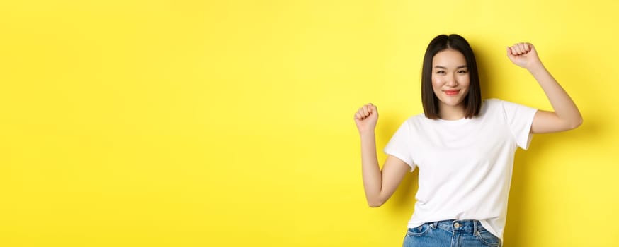 Carefree asian girl dancing and having fun, posing in white t-shirt against yellow background.