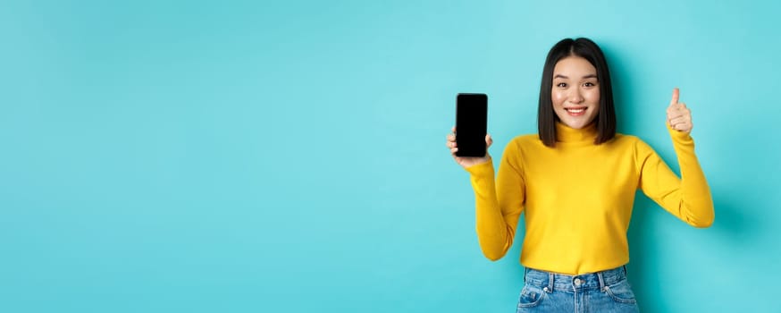 Technology and people concept. Cheerful asian girl in yellow sweater showing blank smartphone screen and thumbs up, demonstrate online offer, standing over blue background.