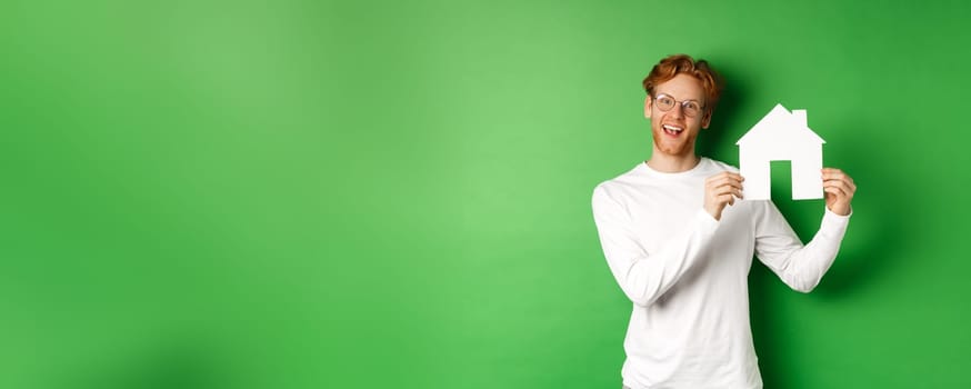 Real estate and buying property concept. Handsome young man with red hair showing house cutout, searching for new home, standing over green background.