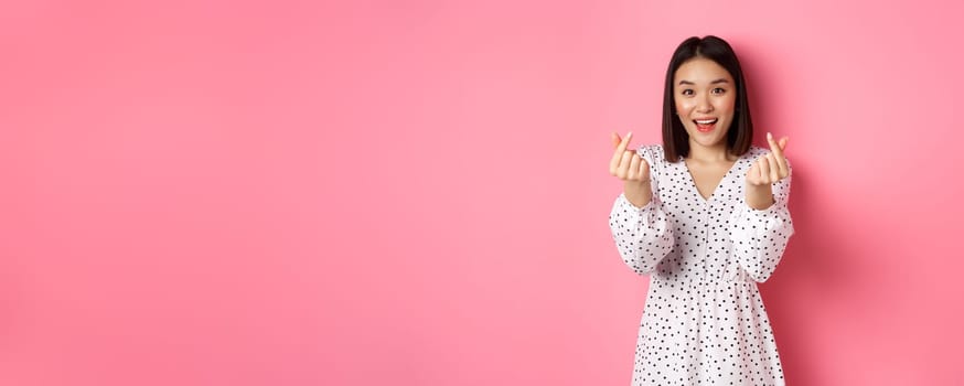 Lovely asian woman in dress showing korean heart signs and smiling, standing on romantic pink background.