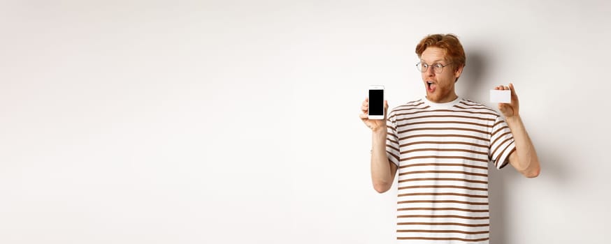 Shopping and finance concept. Amazed young man with red hair showing plastic credit card and smartphone blank screen, staring at display impressed, white background.