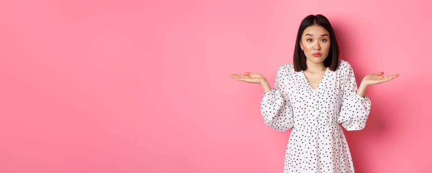 Indecisive cute asian woman shrugging, holding hands spread sideways on copy spaces, dont know, having doubts, standing over pink background.
