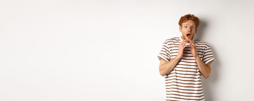 Timid young man with red hair, looking scared, jumping and screaming from fear, standing over white background.