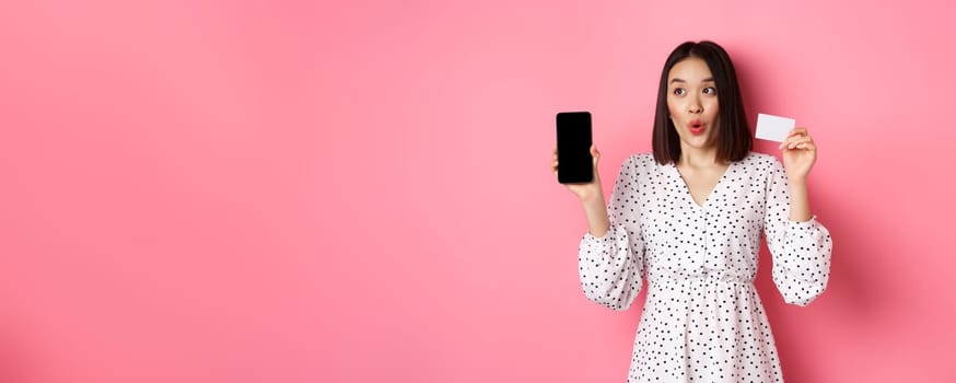 Cute asian woman shopping online, showing bank credit card and mobile screen, smiling and looking left at copy space, standing over pink background.