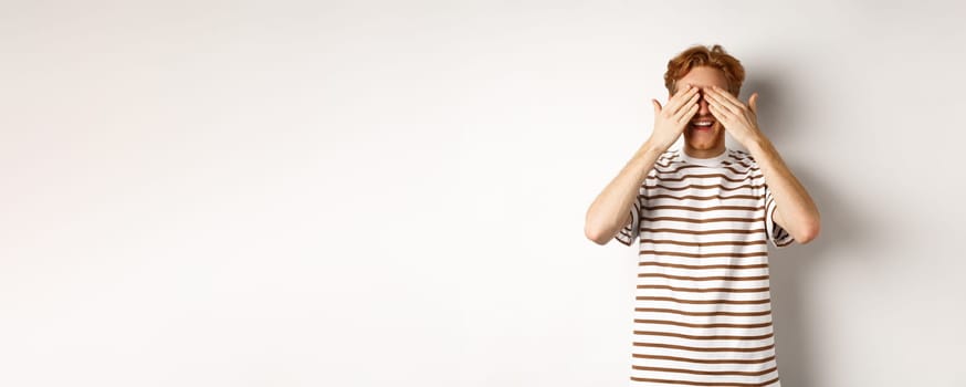 Smiling man cover eyes and waiting for surprise, standing in t-shirt against white background.