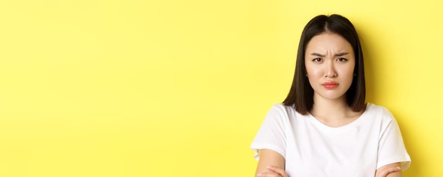 Close up of young asian woman looking angry or disappointed, frowning displeased at camera, standing in white t-shirt, yellow background.