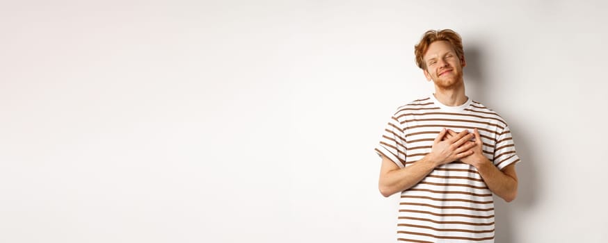 Happy young man with ginger hair appreciate gift, holding hands on heart and smiling, thanking you, standing grateful against white background.