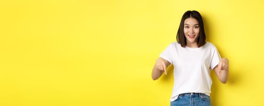 Beauty and fashion concept. Beautiful asian woman in white t-shirt pointing fingers down, demonstrate logo standing over yellow background.