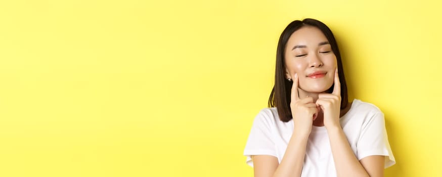 Beauty and skincare. Close up of young asian woman with short dark hair, healthy glowing skin, smiling and touching dimples on cheeks, standing over yellow background.