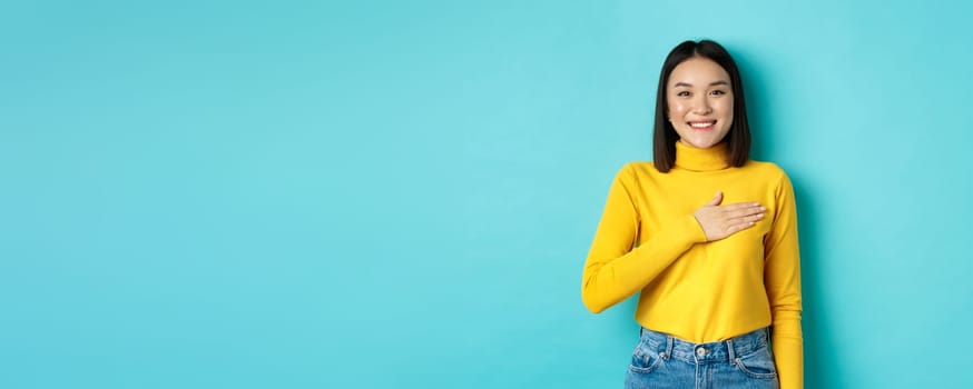 Image of proud smiling asian woman holding hand on heart, showing respect to national anthem, standing over blue background. Copy space