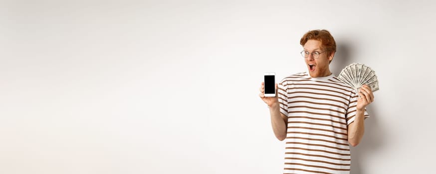 Amazed redhead man showing smartphone app on blank screen and money, winning prize cash online, standing over white background.