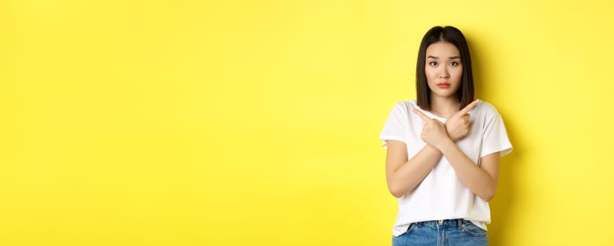 Indecisive asian girl need help with choice, pointing fingers sideways and looking confused, standing over yellow background.