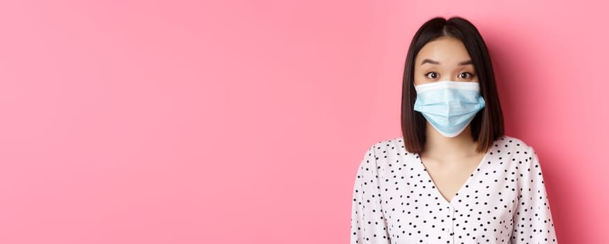 Covid-19, pandemic and lifestyle concept. Surprised asian woman in face mask raising eyebrows, staring at camera amazed, standing against pink background.