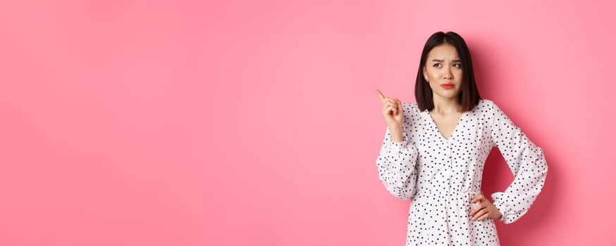 Beautiful asian girl pointing finger at upper left corner, looking confused and doubtful, standing over pink background.