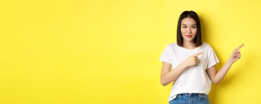 Beauty and fashion concept. Beautiful asian woman in white t-shirt pointing fingers right, demonstrate logo standing over yellow background.