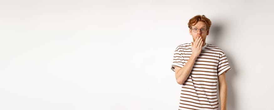 Young man with red hair and glasses hear shocking news or gossiping, cover mouth and staring at camera amazed, white background.