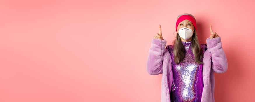 Coronavirus and shopping concept. Fashionable asian elderly woman in disco dress and respirator checking out promo offer, pointing and looking up, pink background.