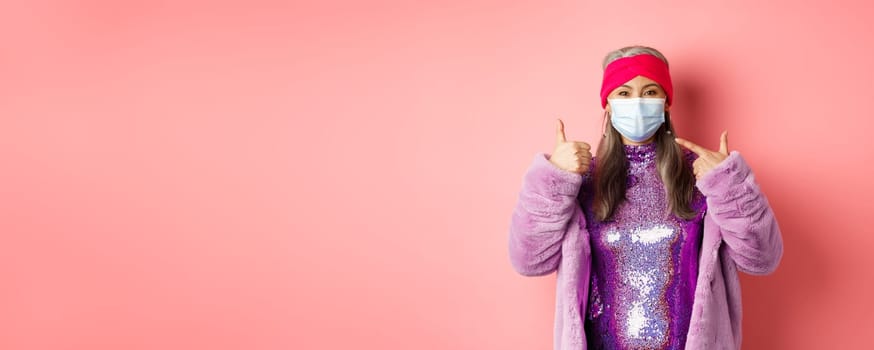 Covid-19, social distancing and fashion concept. Smiling asian senior woman in fashionable clothes and respirator, pointing at face mask and show thumb-up, pink background.