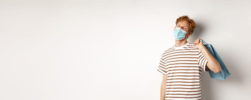 Concept of covid-19 and shopping. Satisfied young man looking pleased after shopping, wearing face mask, holding paper bag and smiling, white background.