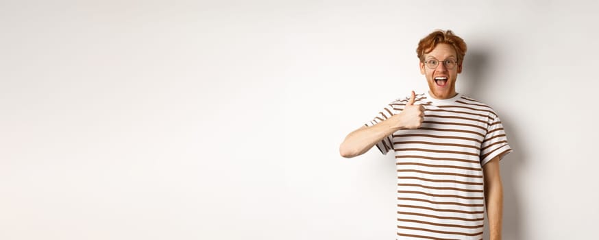 Amazed and fascinated redhead guy checking out something awesome, showing thumb-up and scream yes with joy, standing over white background.