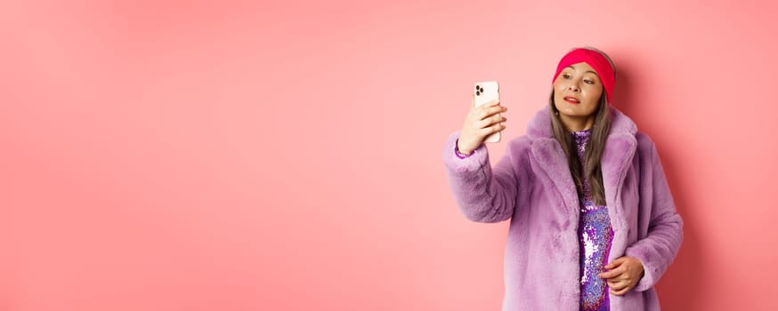 Fashion concept. Stylish asian senior female taking selfie on smartphone, posing in purple faux fur coat and party dress, standing over pink background.