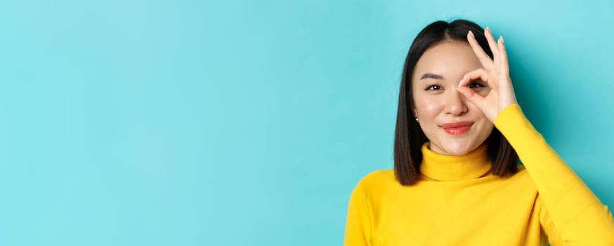 Beauty and makeup concept. Close up of carefree asian girl showing OK sign on eye and smiling, looking happy at camera, standing over blue background.