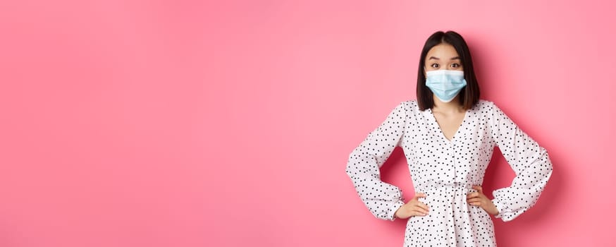 Covid-19, quarantine and lifestyle concept. Cute korean woman in dress and face mask using preventive measures from coronavirus, standing over pink background.