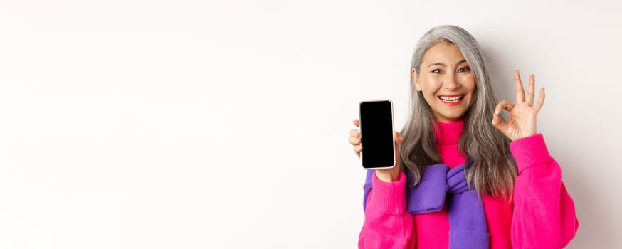 Online shopping. Fashionable asian senior woman showing blank smartphone screen and OK sign, smiling pleased, recommending app, white background.