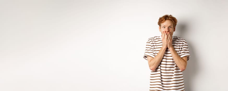 Surprised and excited man with curly red hair checking out special promo offer,staring fascinated and amazed, covering mouth with hands, standing over white background.