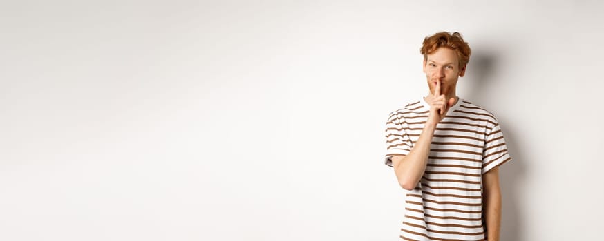 Young man student with red hair shushing, telling a secret and smiling, standing over white background.