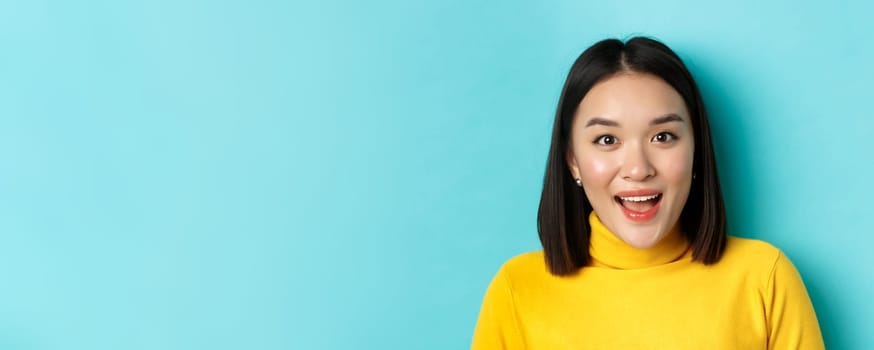 Beauty and skincare concept. Close up of surprised asian female raising eyebrows and look amused at camera, look at something interesting, standing over blue background.