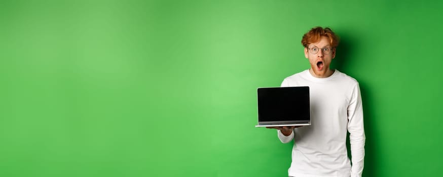 Impressed redhead guy in glasses drop jaw after seeing online promo, showing laptop screen and staring at camera with disbelief, standing over green background.