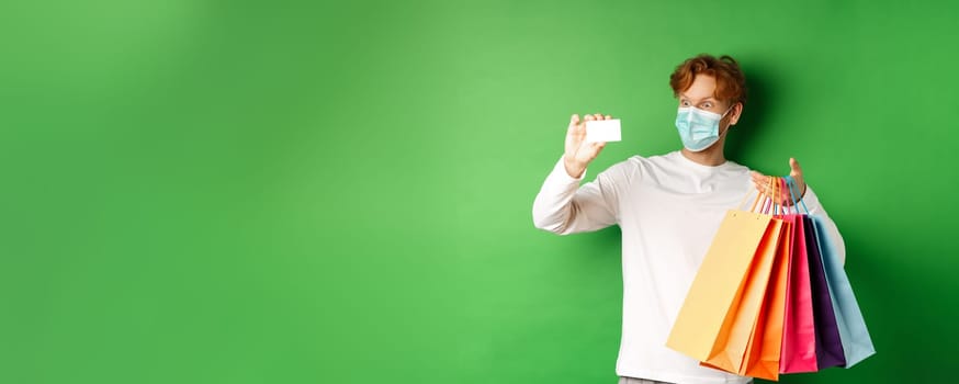 Handsome young man in medical mask, showing plastic credit card and shopping bags with items purchased with discount, green background.