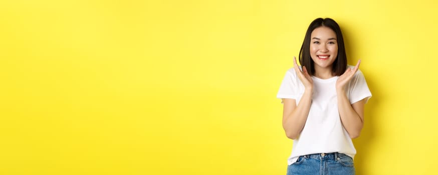 Beauty and fashion concept. Excited asian woman clap hands and smiling happy at camera, standing in white t-shirt against yellow background.