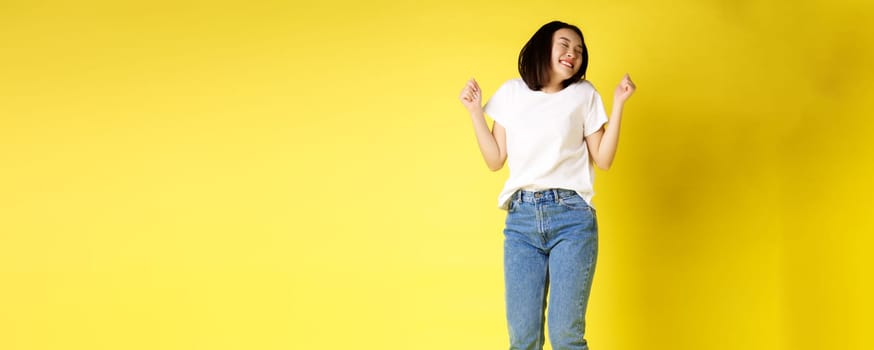 Full size shot of carefree asian woman jumping and dancing, having fun, posing in jeans and white t-shirt over yellow background.