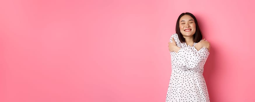 Beautiful asian woman feeling romantic, hugging herself, close eyes and smiling with dreamy face, standing happy over pink background.