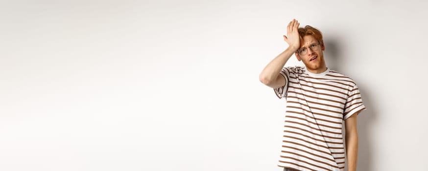 Tired and distressed redhead male student slap forehead, making facepalm and staring at camera exhausted, standing over white background.