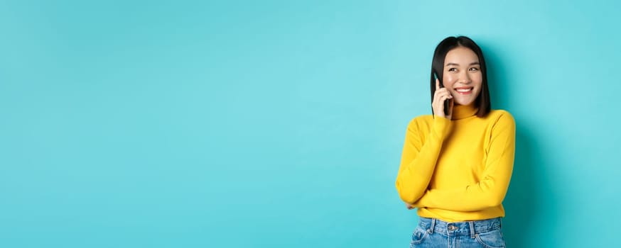 Attractive korean woman in yellow sweater, having conversation and smiling, talking on mobile phone, looking left at copy space, standing over blue background.