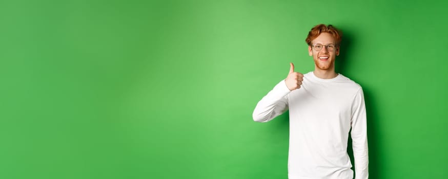 Handsome young man in glasses with red hair, showing thumbs-up and smiling satisfied, praising good eyewear store, standing over green background.