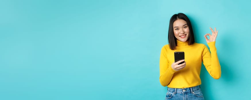 E-commerce and online shopping concept. Portrait of asian woman showing OK sign and using mobile phone, praise app, standing over blue background.