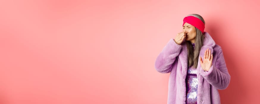 Disgusted asian mature woman in trendy purple winter coat and dress, shut nose and showing stop, refusal gesture, smell something disgusting, standing over pink background.