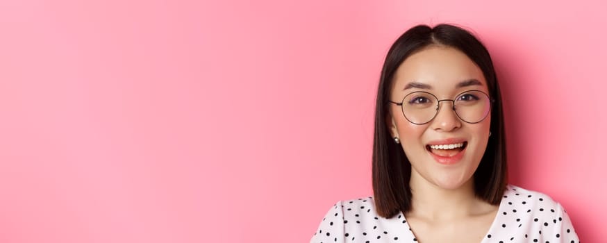 Beauty and lifestyle concept. Close-up of cute asian female model wearing trendy glasses, smiling happy at camera, standing on pink background.