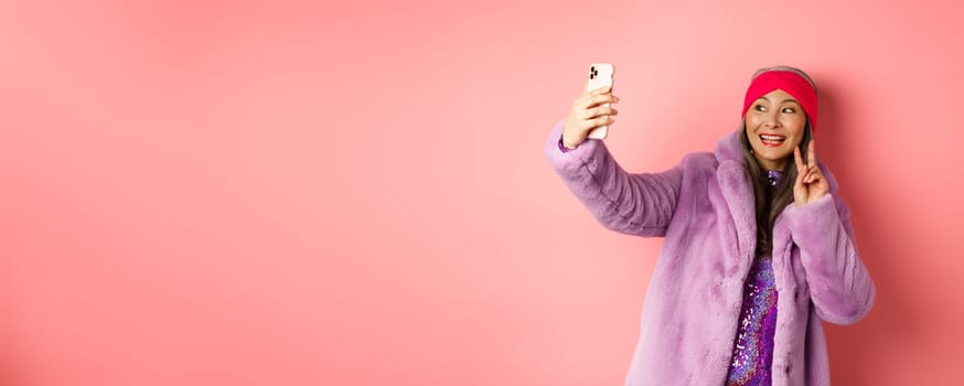 Fashion concept. Cool and cheerful asian senior woman taking selfie with peace sign, wearing trendy faux fur coat and party dress, standing over pink background.