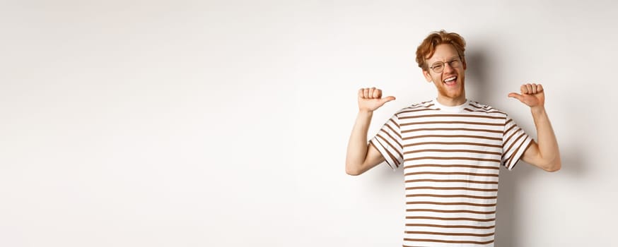 Cheerful and funny redhead man with beard, wearing glasses, laughing and pointing at himself, standing over white background.
