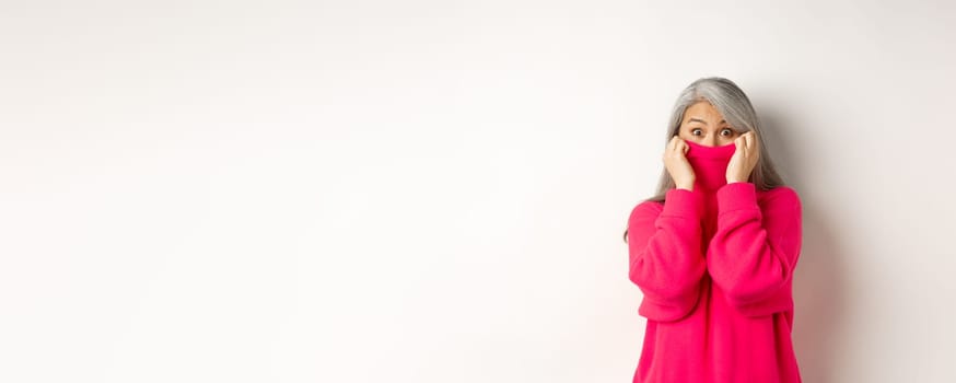 Portrait of funny asian grandmother hiding face in sweater collar, peeking at camera silly, standing over white background.