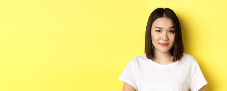 Close up of beautiful asian woman with casual makeup, raising eyebrow and looking intrigued at camera, standing curious over yellow background.