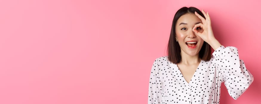 Beauty and lifestyle concept. Beautiful asian girl looking through okay sign and smiling amazed, standing against pink background.