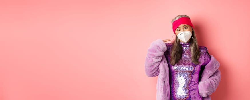 Covid-19, pandemic and fashion concept. Asia senior woman in stylish glittering dress complaining on face mask, pointing at respirator and frowning, pink background.