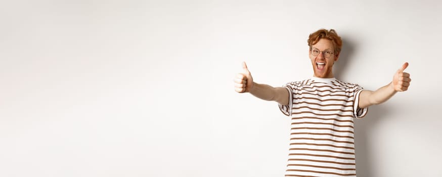 Happy redhead man scream yes and showing thumbs-up, approve and praise excellent company, standing over white background.